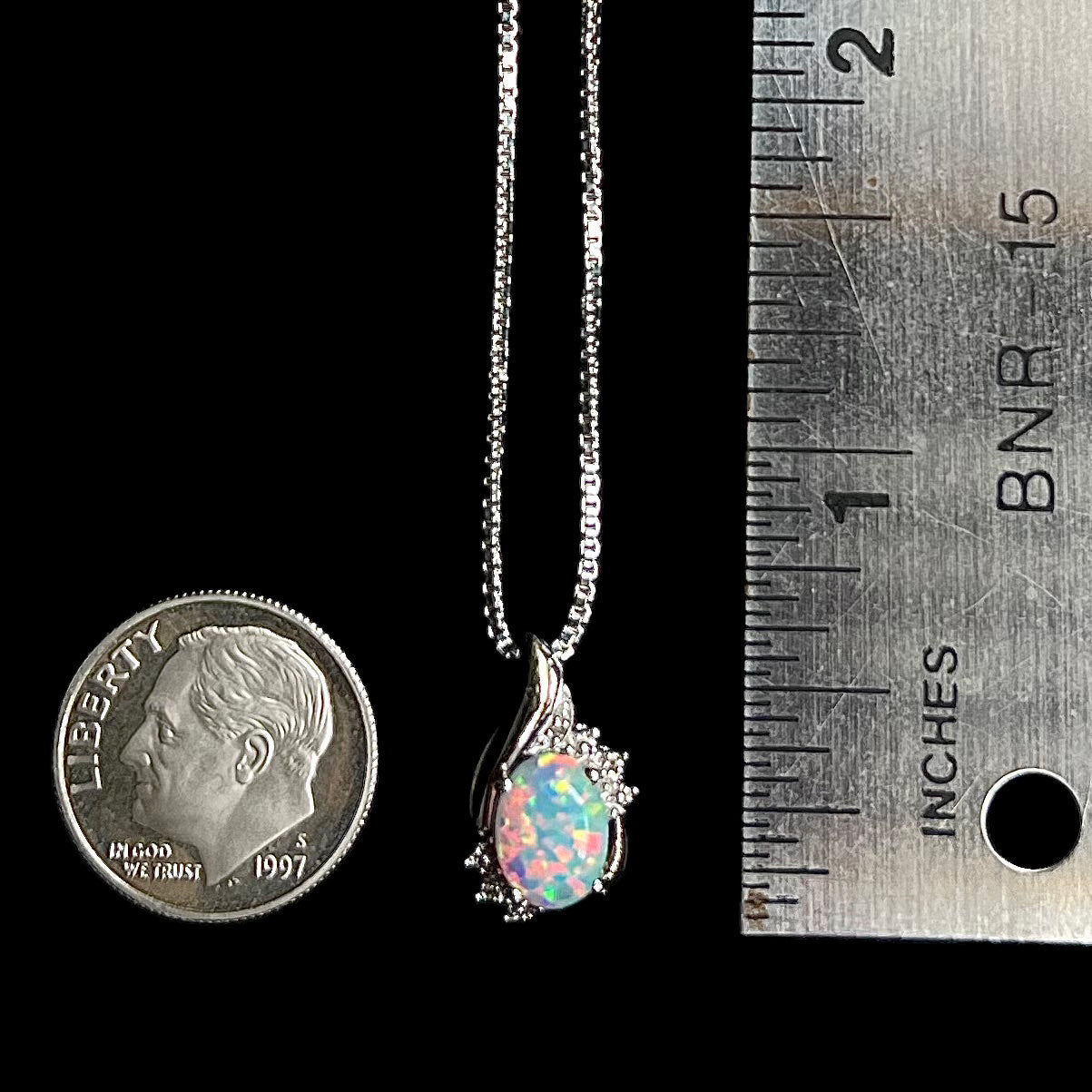 Lab-created opal and diamond pendant in sterling silver measuring half of an inch long on a chain next to a ruler and dime coin for scale.