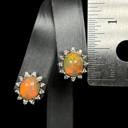 Half inch-long Ethiopian opal and diamond halo stud earrings on earring display next to ruler