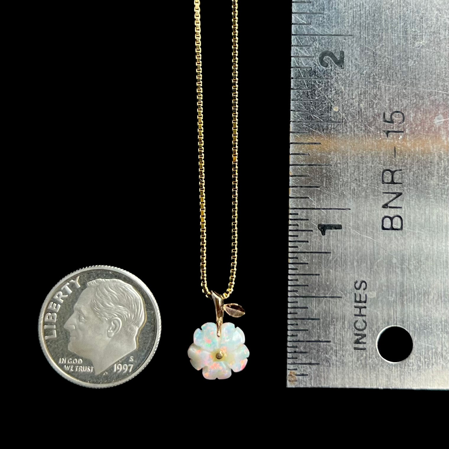 An opal flower pendant that measures half of an inch long shown next to a ruler and dime coin against a black background.