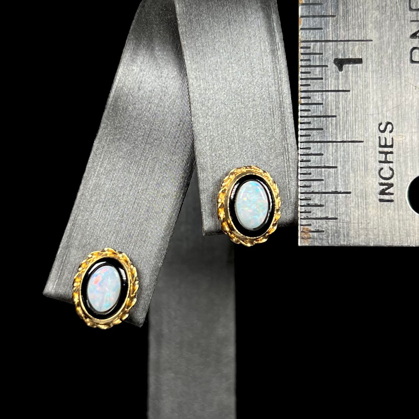 Half inch-long gold opal in onyx stud earrings on an earring display next to ruler.