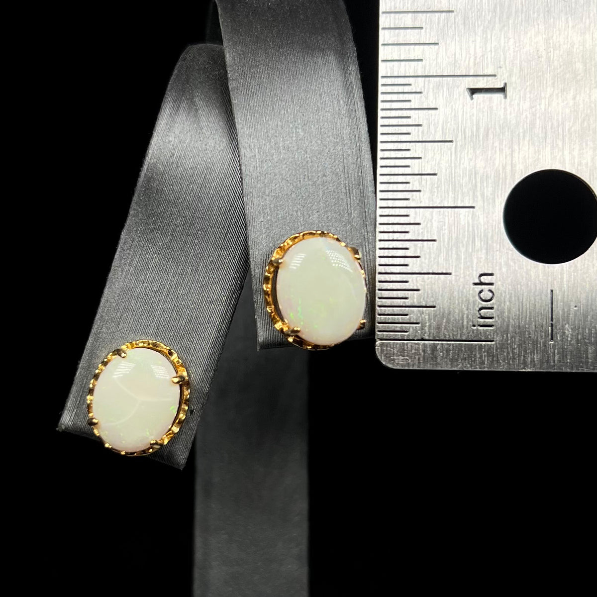 Half inch-long oval shaped opal stud earrings on earring display next to ruler
