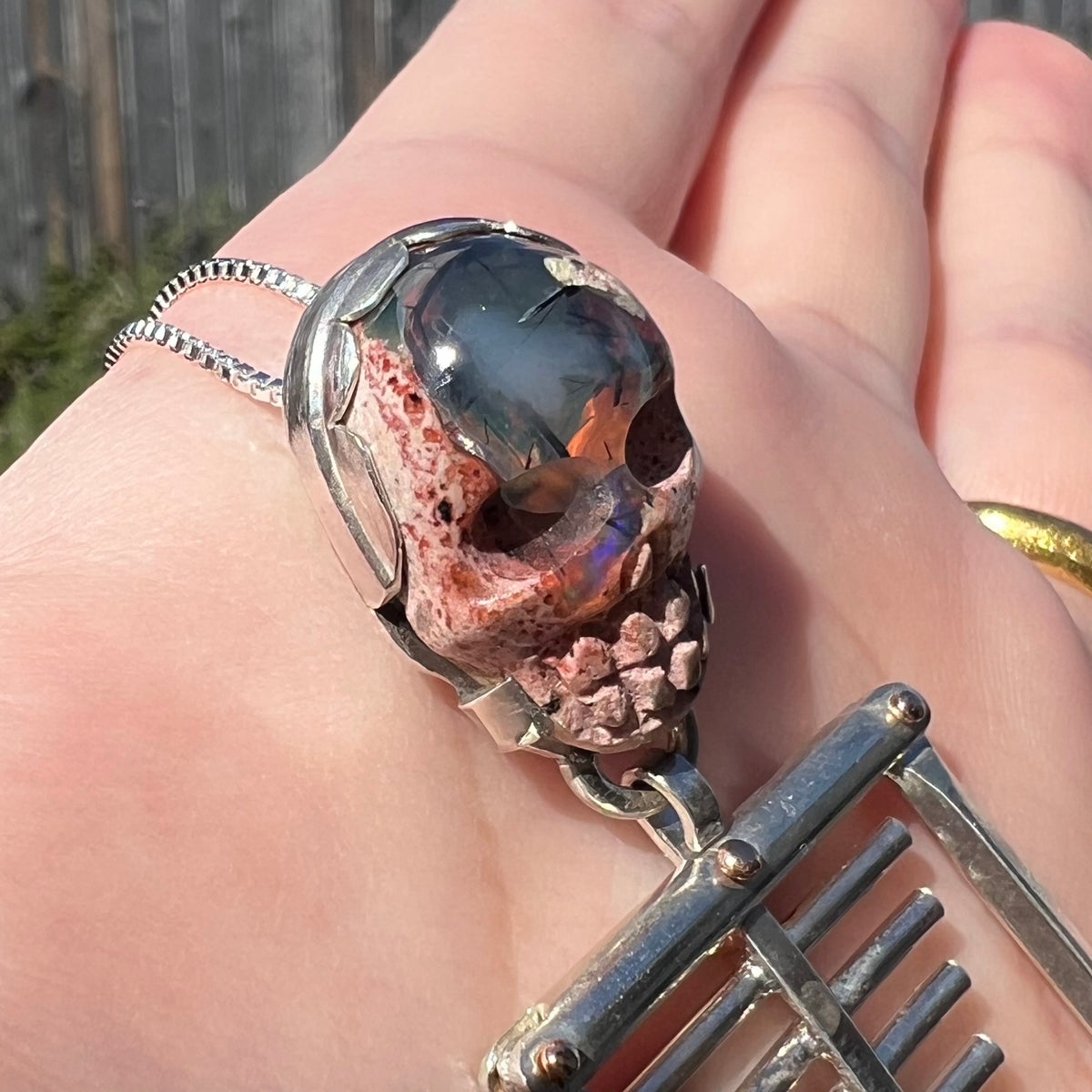 Angled close-up of Halloween-inspired  sterling silver skeleton pendant featuring carved cantera matrix opal skull, held in natural sunlight