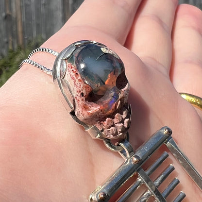 Angled close-up of Halloween-inspired  sterling silver skeleton pendant featuring carved cantera matrix opal skull, held in natural sunlight