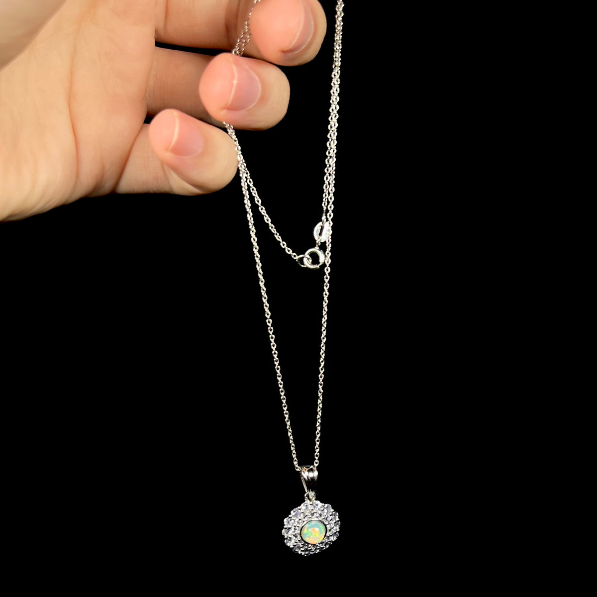Hand dangling a sterling silver Ethiopian opal and tanzanite halo women's pendant from its cable-style chain.