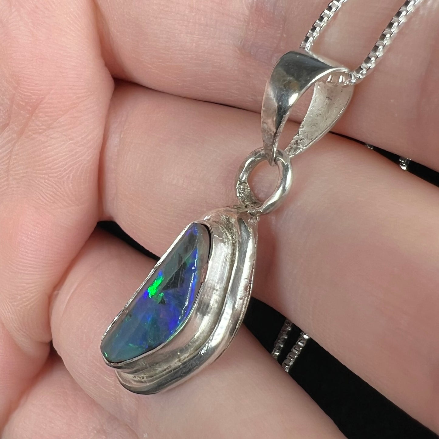 Angled close-up of a handmade sterling silver black opal pendant.  Tooling marks are visible in the silver, as is a green flash in the opal.