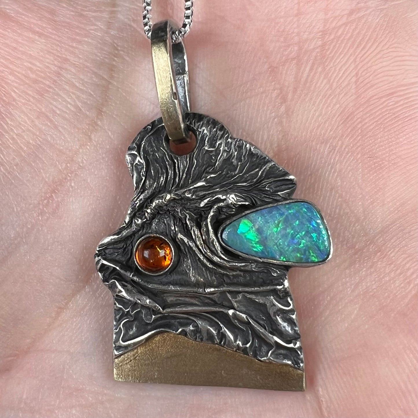 Close-up of a handmade sterling silver and 18k gold men's opal and amber pendant, shown held in a hand.