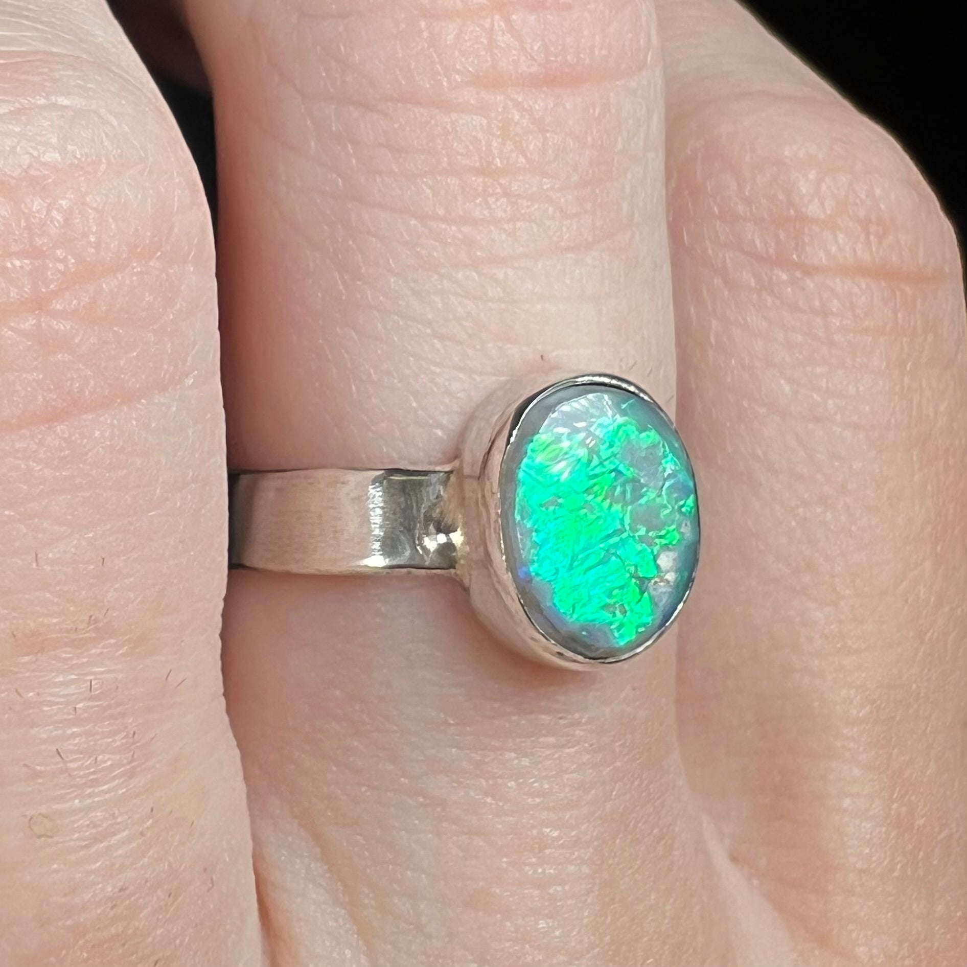 Finger wearing a handmade sterling silver Lightning Ridge opal solitaire ring with a teal flash across the opal's surface, shown from an angle.