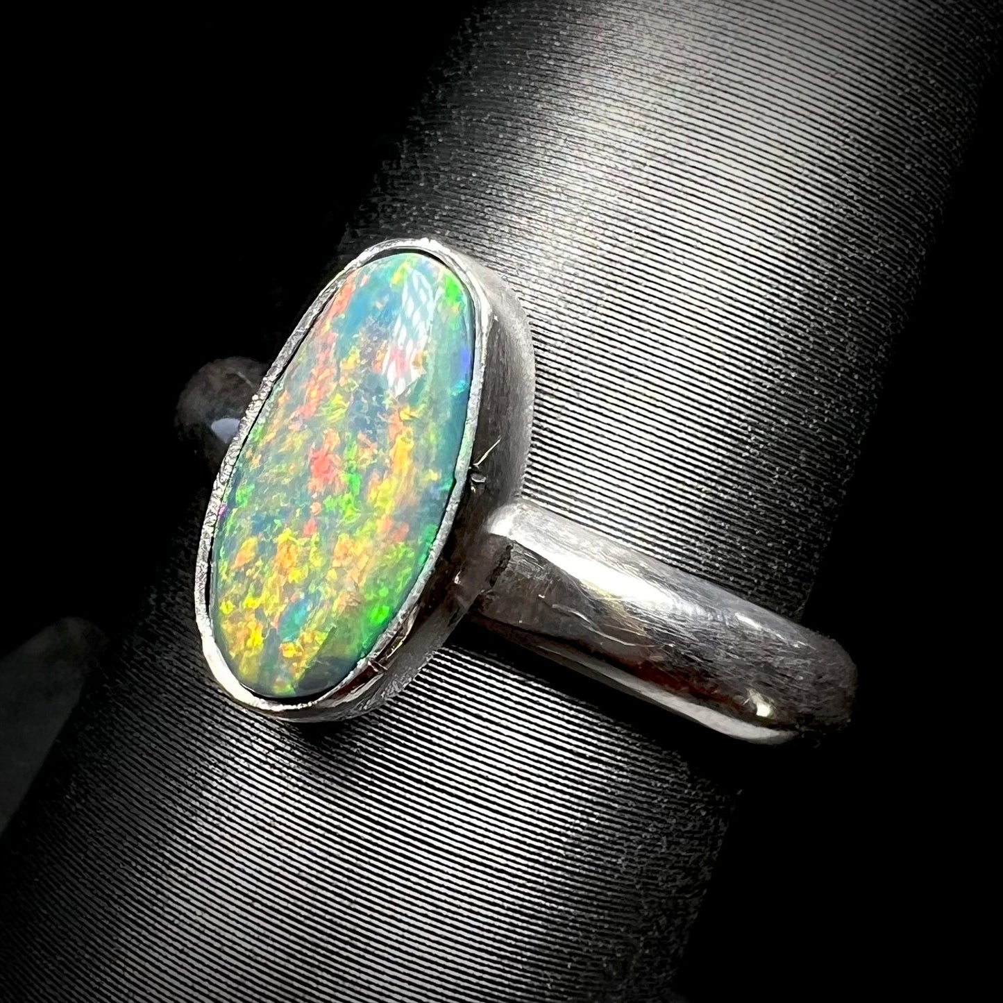 Angled close-up of a handmade sterling silver Australian dark opal solitaire ring shown on a ring display.  Tooling marks are seen on the opal's bezel setting.