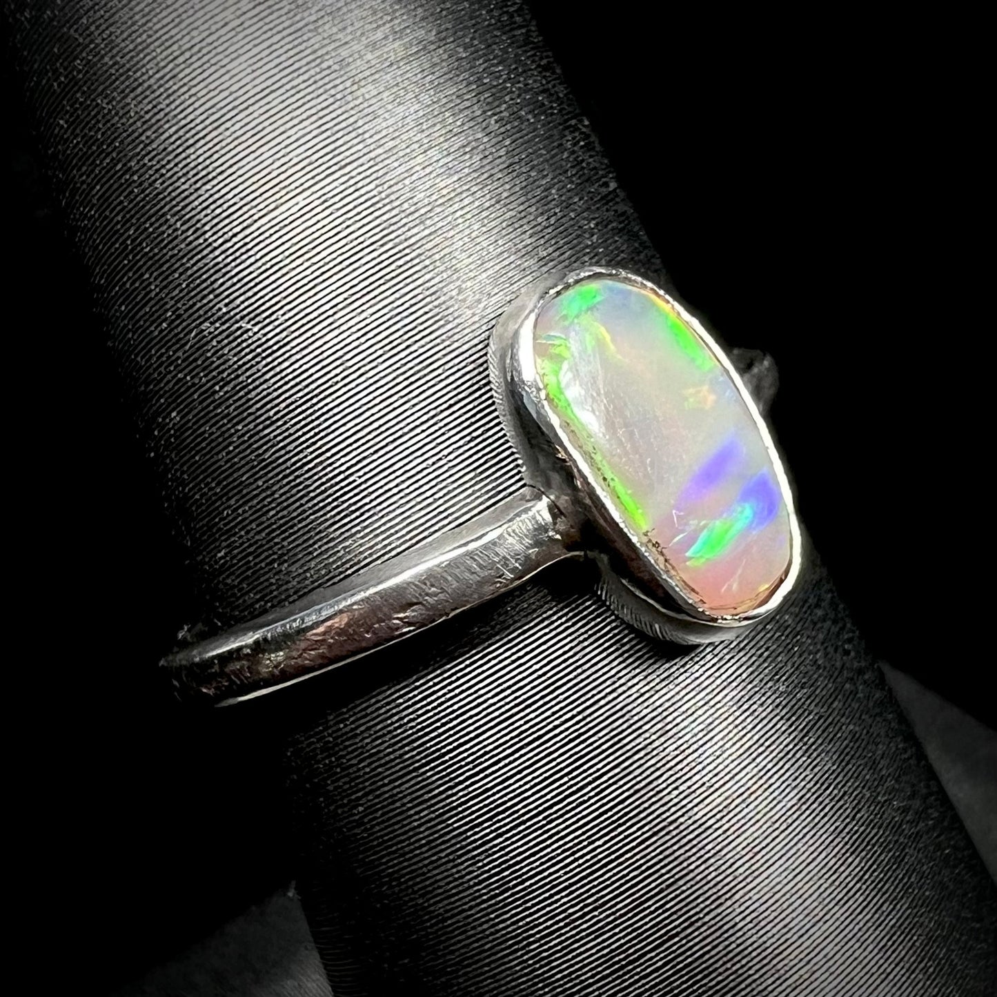 Angled close-up of a handmade sterling silver opal solitaire ring on a ring display.  The opal has purple and green play of color.