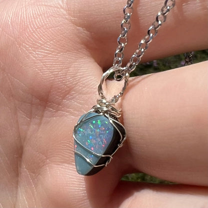 Angled close-up of a handmade sterling silver wire wrapped opal doublet women's pendant, shown in a hand under natural sunlight.