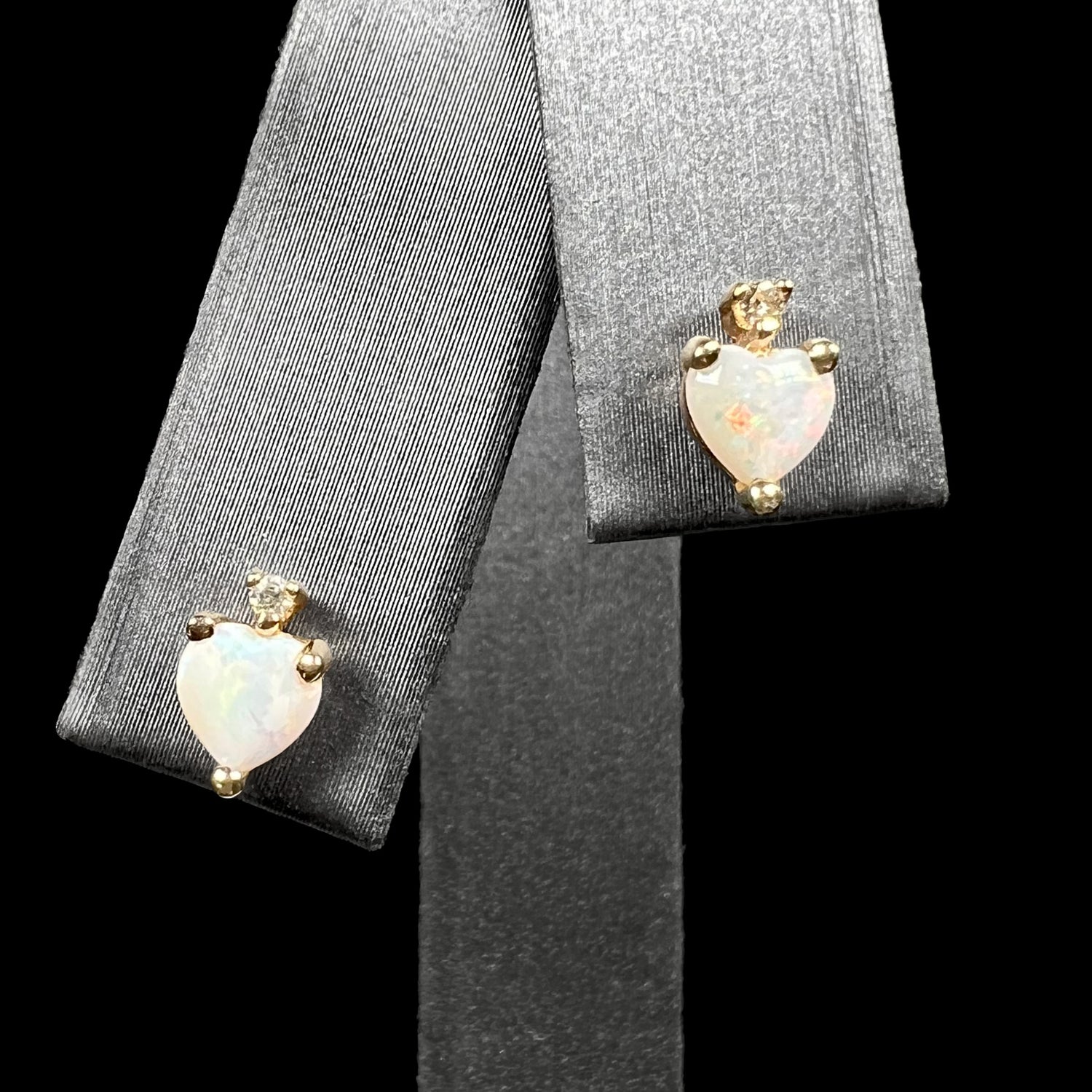 A pair of heart shaped Australian opal and diamond stud earrings in 14k yellow gold, shown on an earring display against black background.