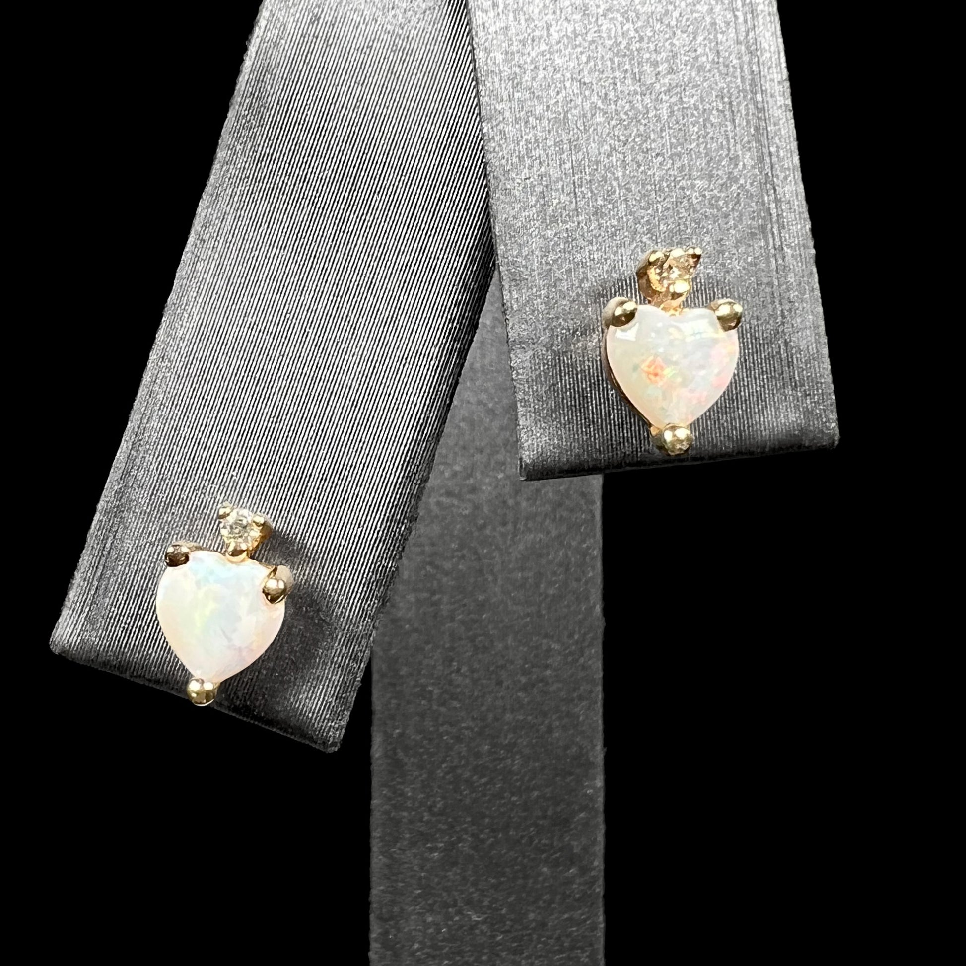 A pair of heart shaped Australian opal and diamond stud earrings in 14k yellow gold, shown on an earring display against black background.