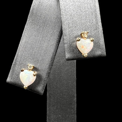A pair of heart shaped Australian opal and diamond stud earrings in 14k yellow gold, shown on an earring display against black background.