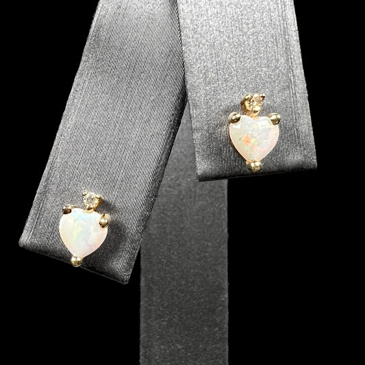 A pair of heart shaped Australian opal and diamond stud earrings in 14k yellow gold, shown on an earring display against black background.