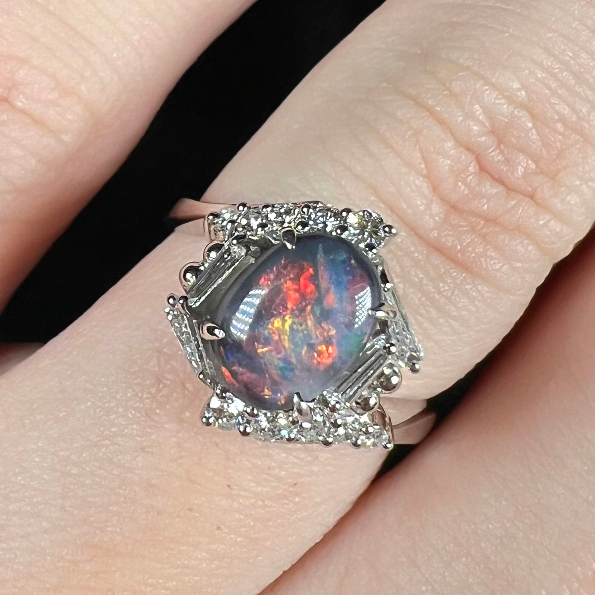 Close-up of N5 dark opal ring featuring hexagonal halo design set with round and tapered baguette cut diamonds, worn on finger.