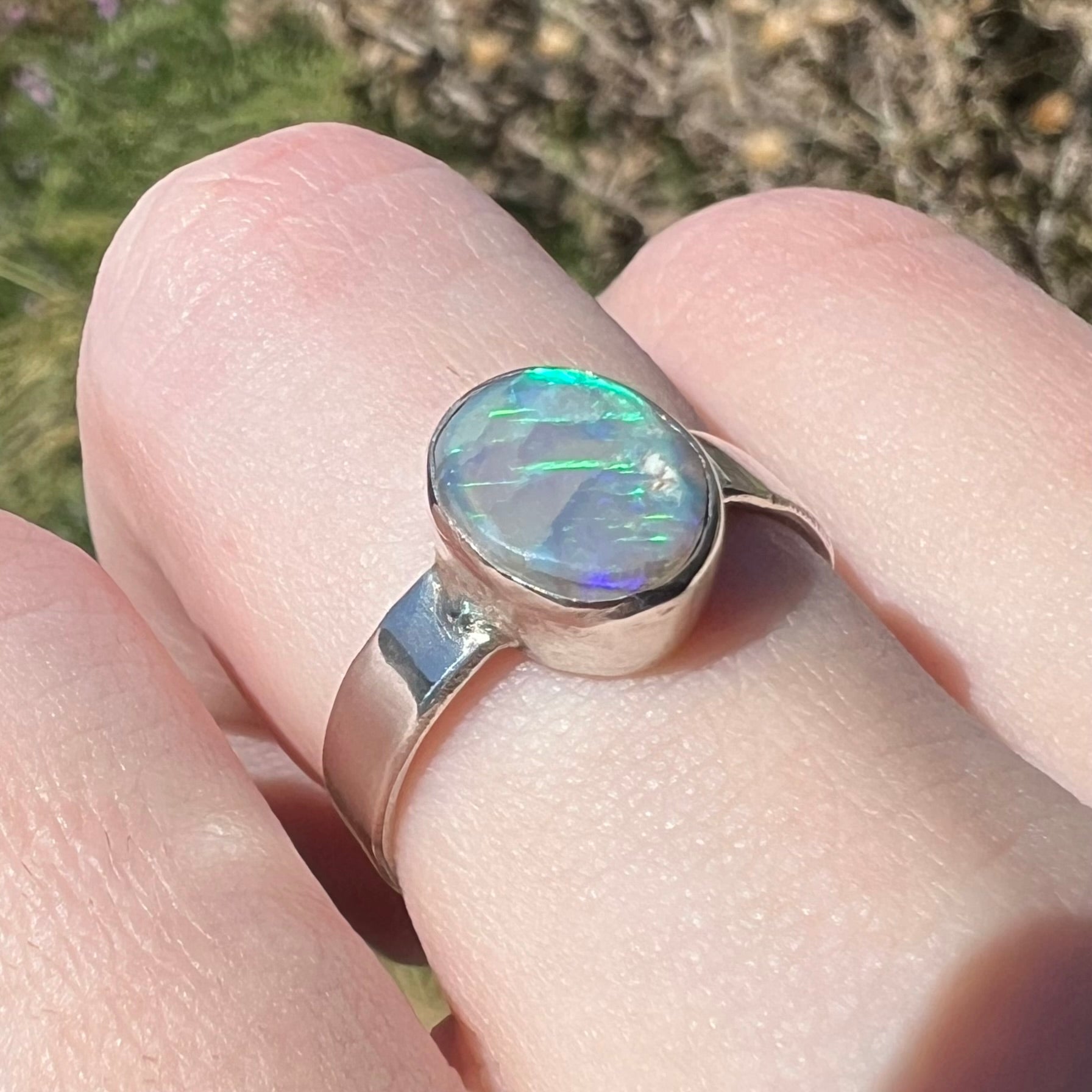 A sterling silver highly-directional Australian opal ring, worn on a finger in sunlight and shown from an angle; the opal has soft green and blue streaks of color.