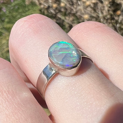 A sterling silver highly-directional Australian opal ring, worn on a finger in sunlight and shown from an angle; the opal has soft green and blue streaks of color.