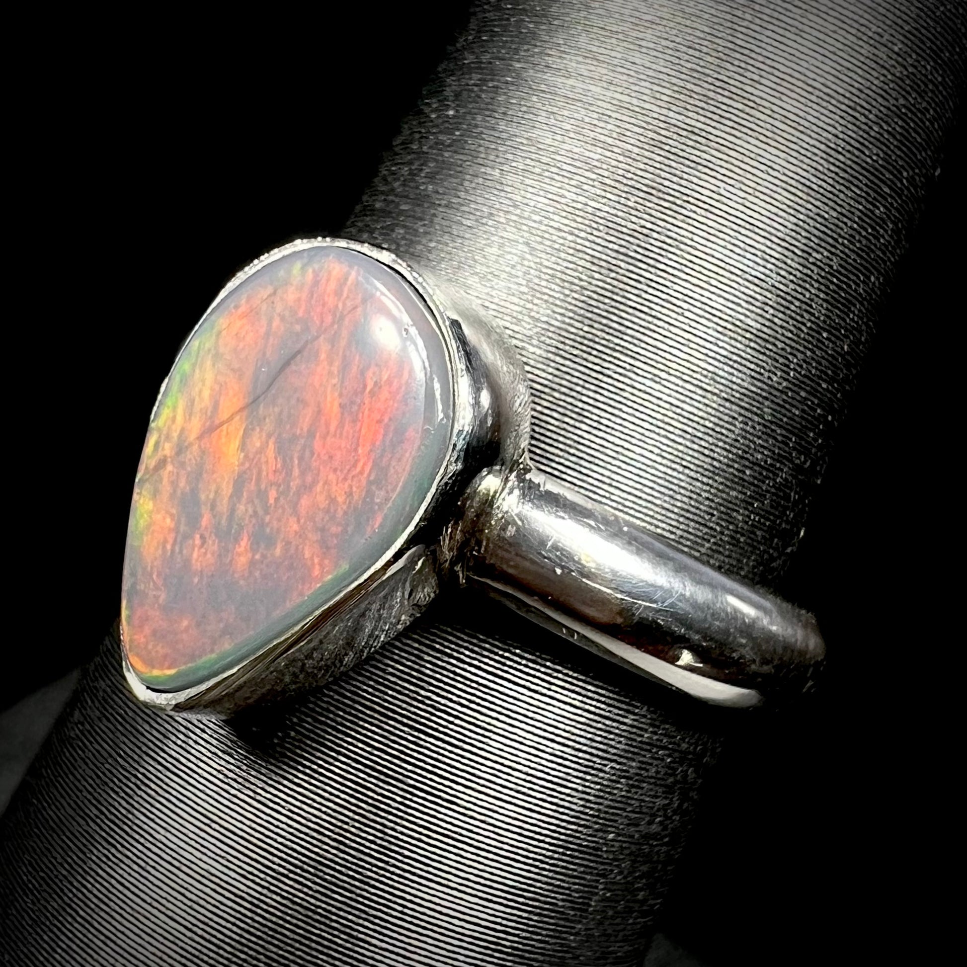 Angled close-up of a natural dark opal ring in sterling silver with highly reddish, orange, and green flashes of color, shown on a ring display.