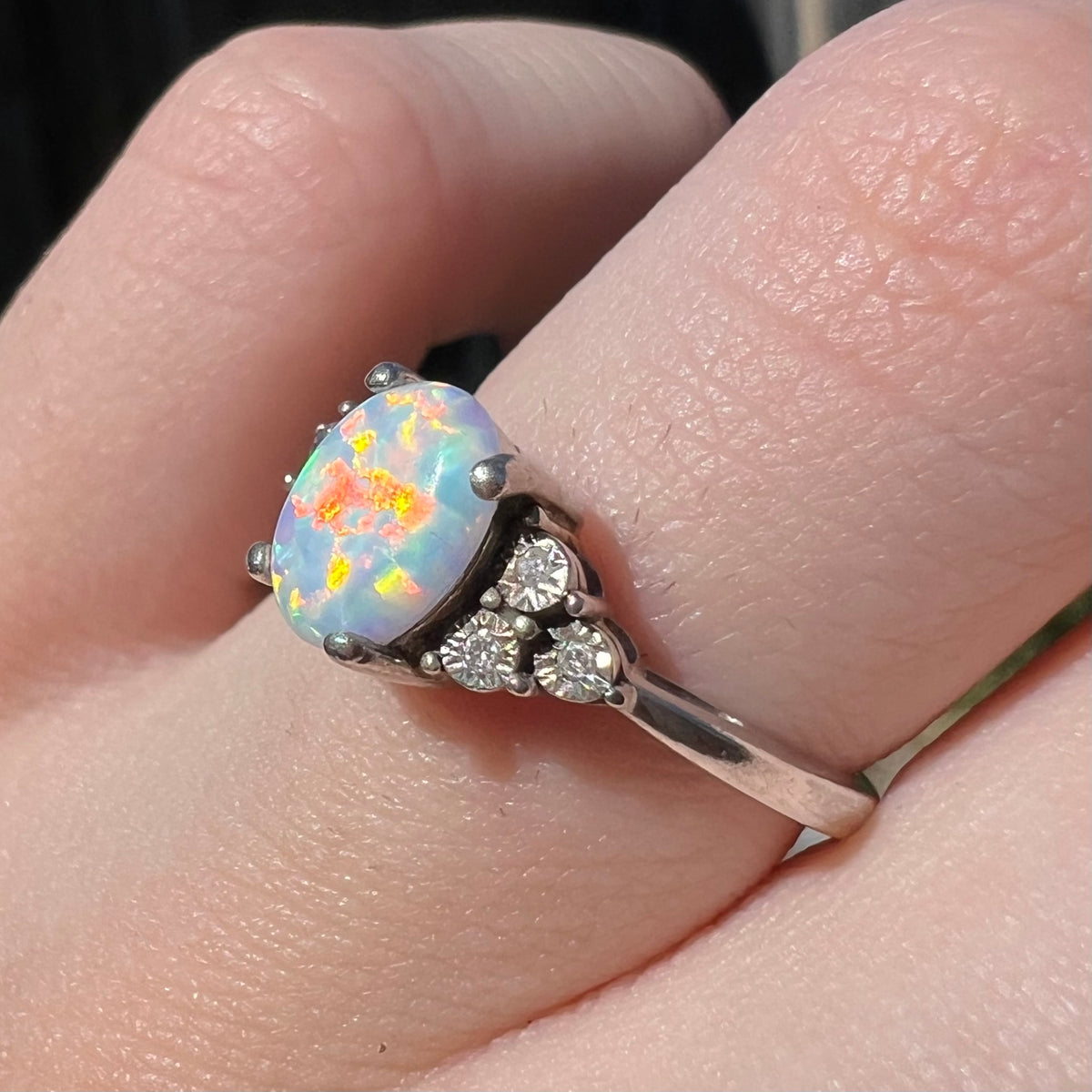Red and orange play-of-color seen in lab-created opal ring, worn on finger in sunlight.  Three illusion-set diamonds mounted in the ring's shoulders are in view.