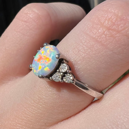 Red and orange play-of-color seen in lab-created opal ring, worn on finger in sunlight.  Three illusion-set diamonds mounted in the ring's shoulders are in view.
