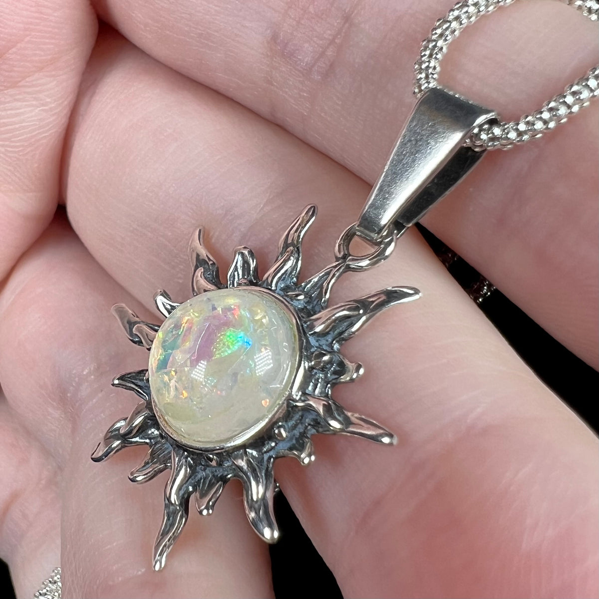 A silver sunburst pendant necklace with a central iridescent imitation opal glass gemstone, held in a person's hand and shown from an angled macro view.
