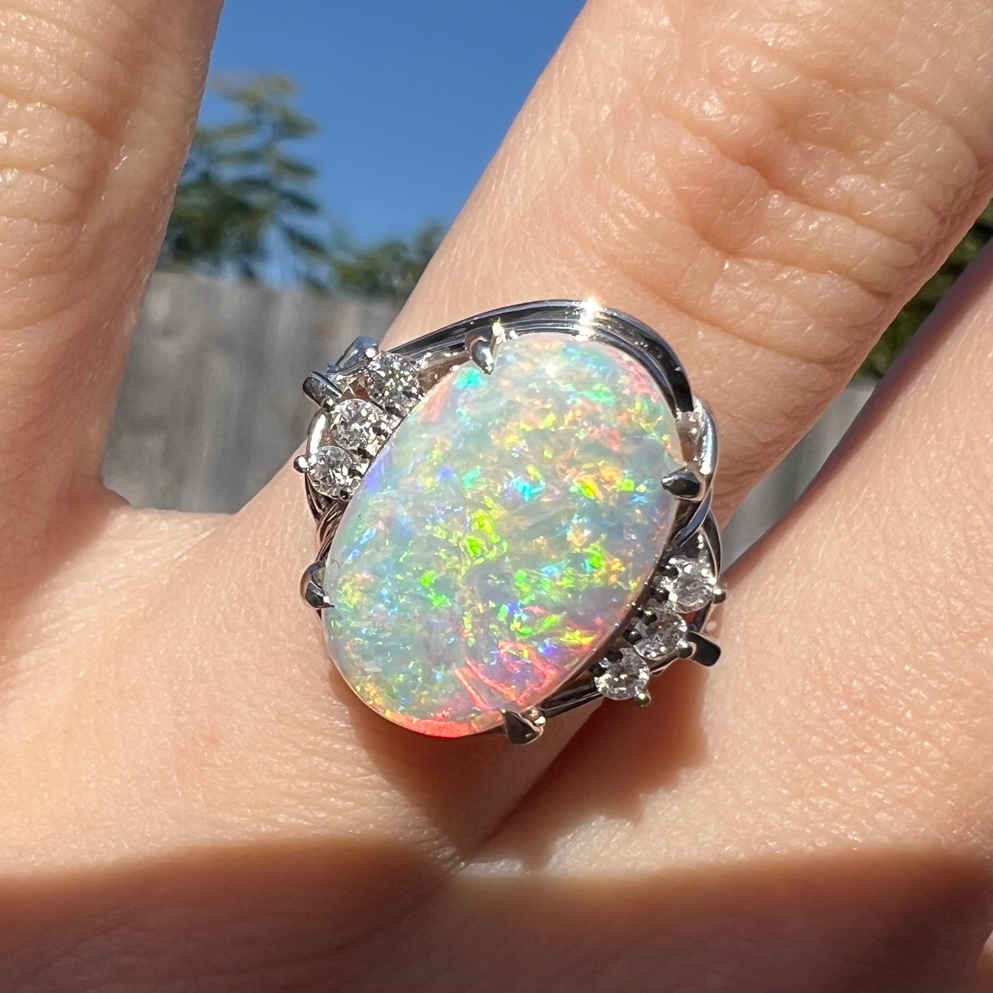 A Japanese opal and diamond ring worn on a hand in sunlight, showcasing the opal's green and orange color play.