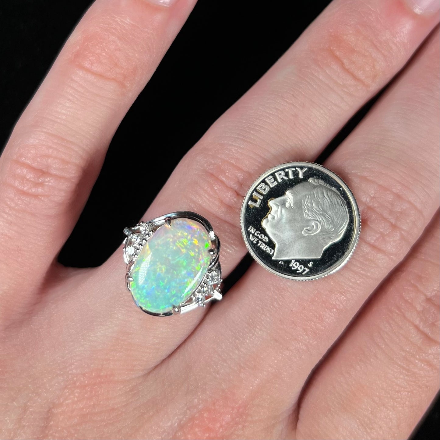 A platinum Japanese opal and diamond ring on a hand, being compared with a dime for scale.