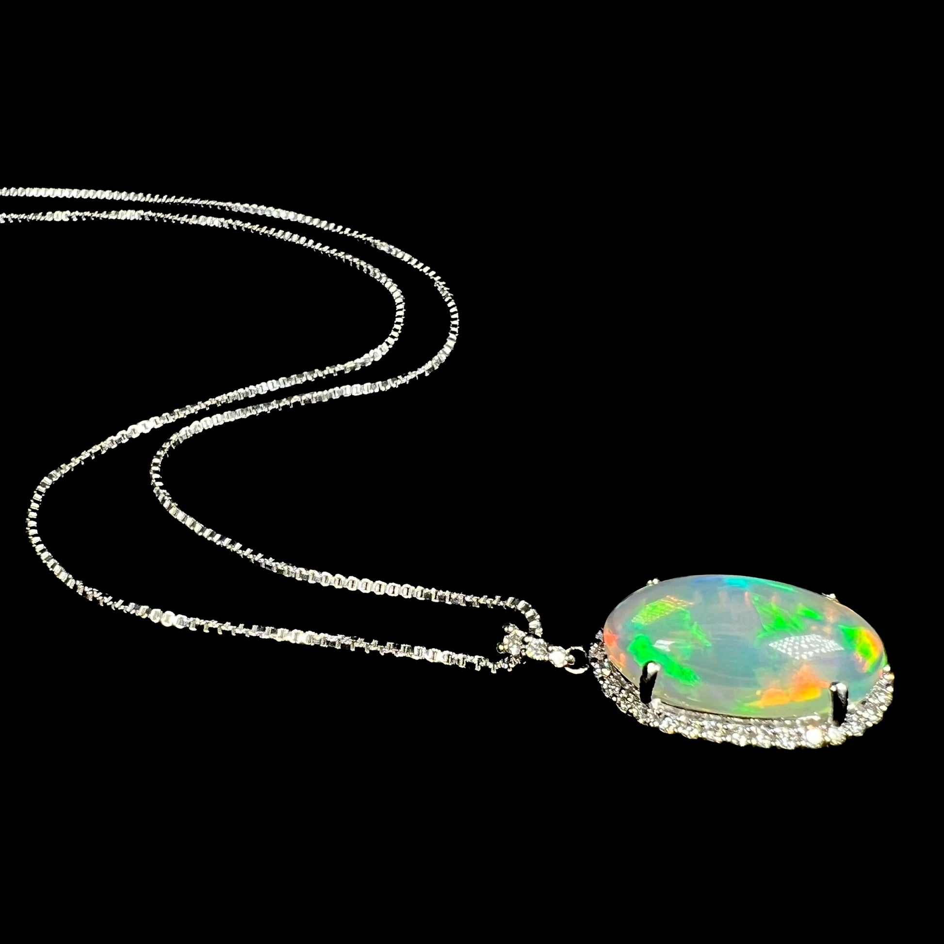Angled view of a jelly crystal opal and diamond halo necklace in platinum, laying on a black background.  The opal has green and red colors.