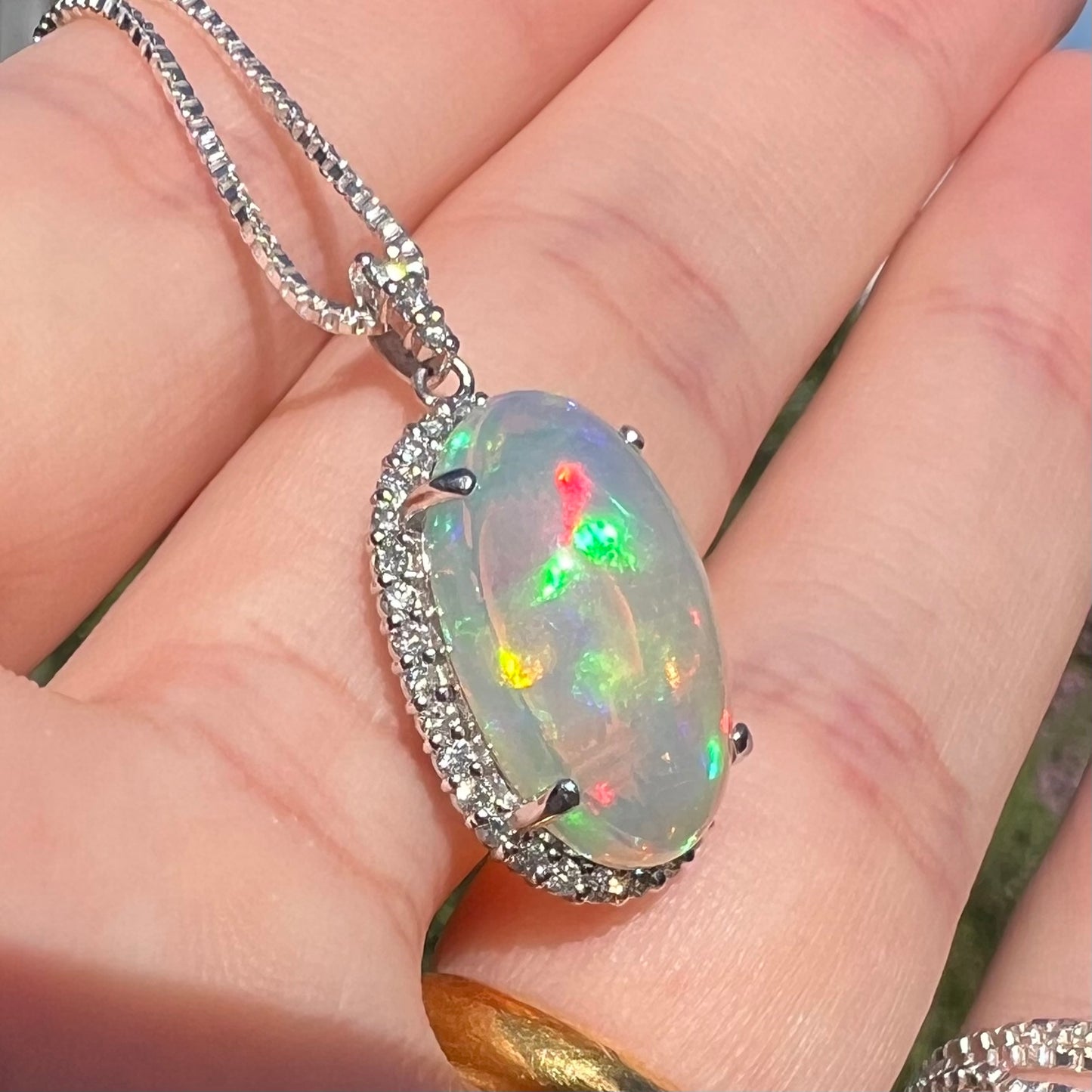 Angled view of a jelly crystal opal and diamond halo pendant, shown at an angle in a hand under sunlight.  The opal has green and red flashes of color.