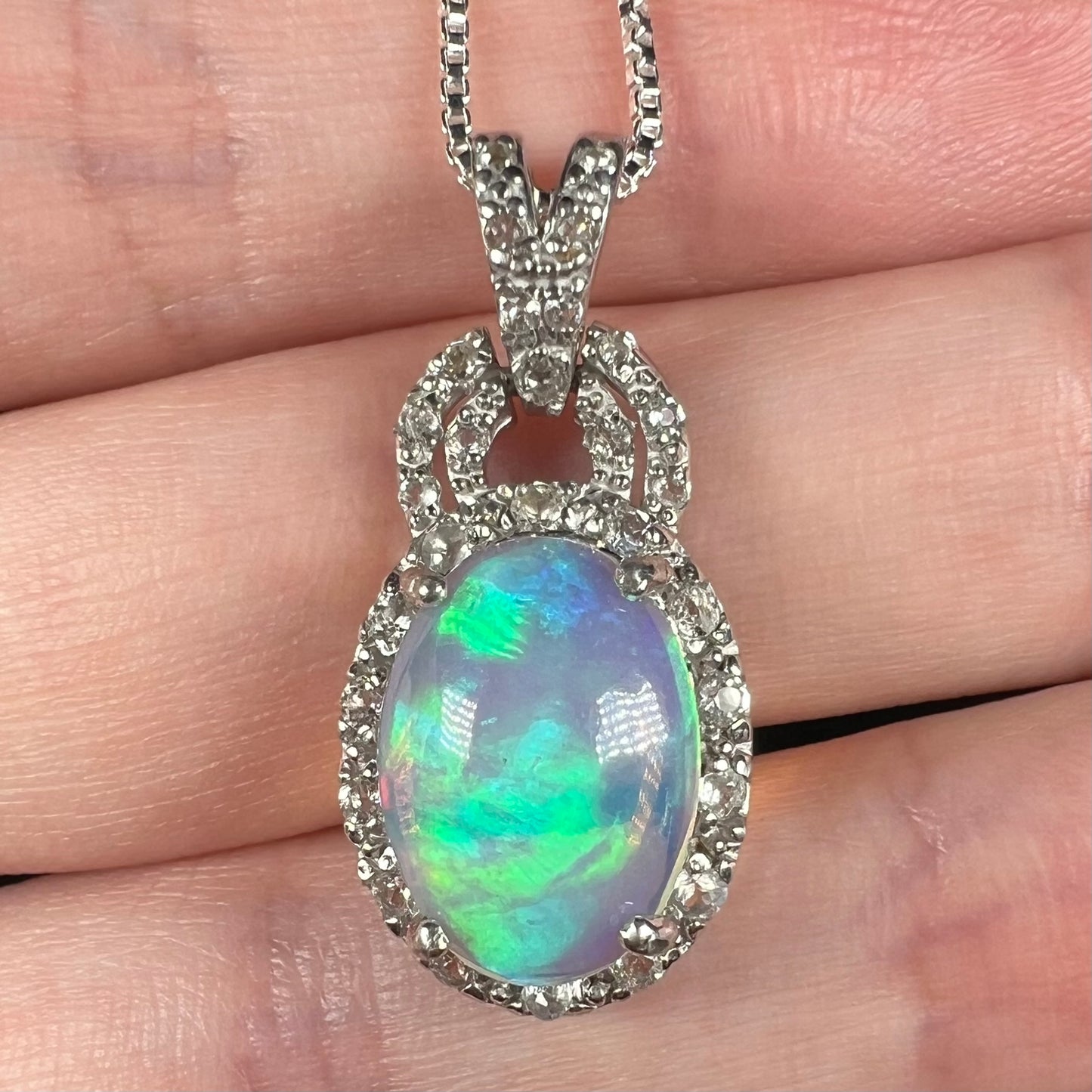 Close-up of Ethiopian jelly opal pendant with blue and green flashes in white topaz halo design, held in hand