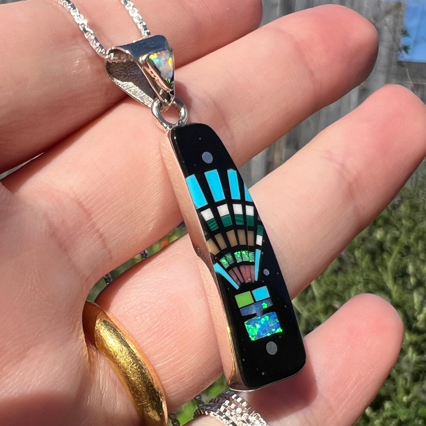 Lab-created opal and black onyx pendant with kachina motif shown close-up from angle in hand under natural sunlight.