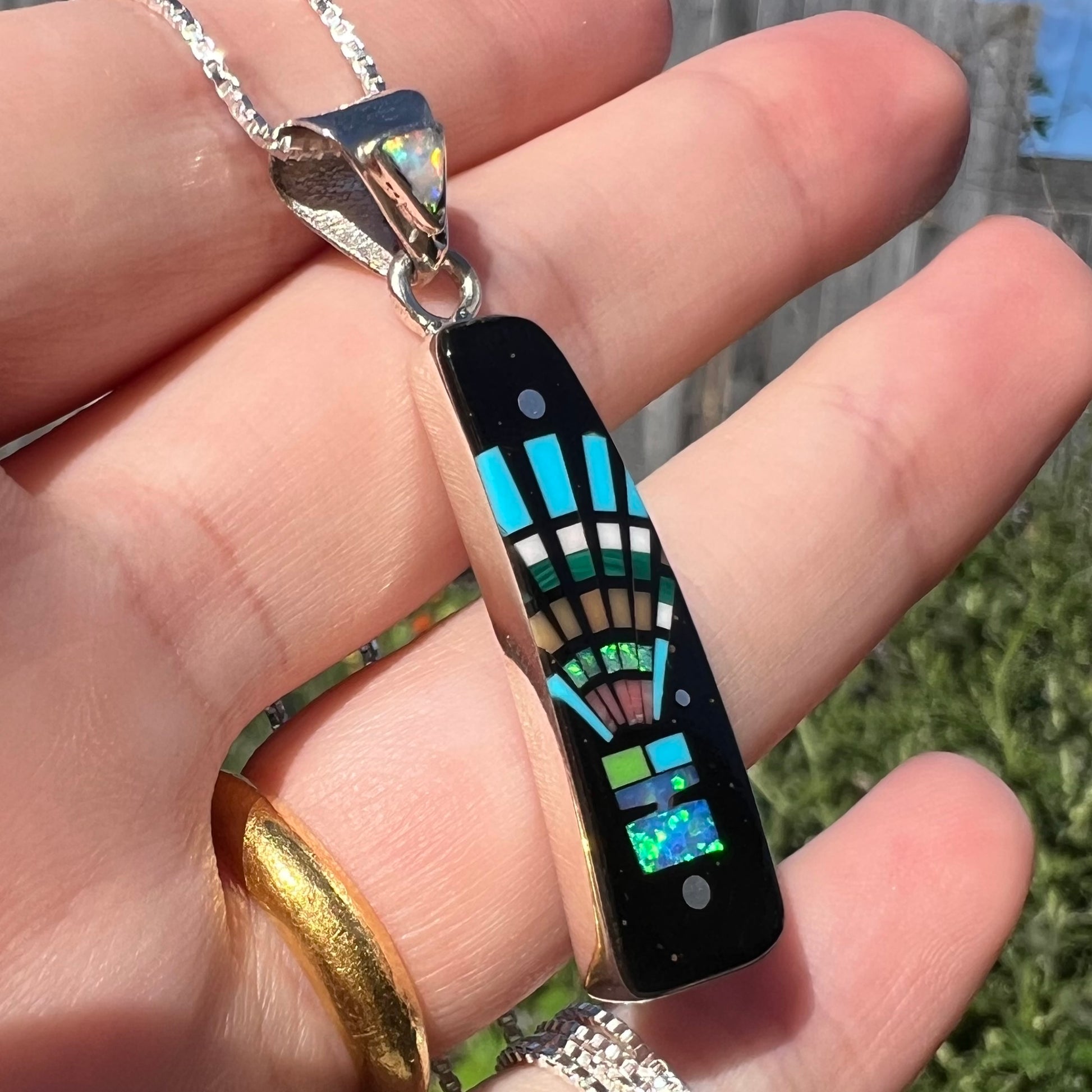 Lab-created opal and black onyx pendant with kachina motif shown close-up from angle in hand under natural sunlight.