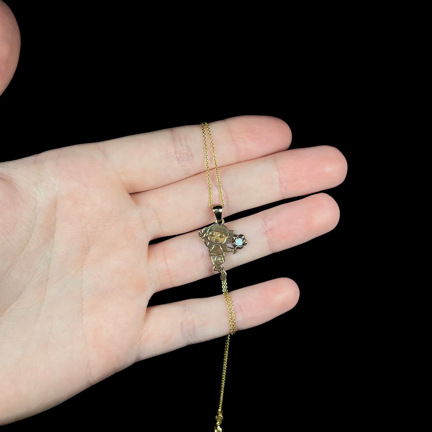 Hand holding a kids gold-tone sterling silver Dora the Explorer opal necklace in front of a black background under cool studio lighting.