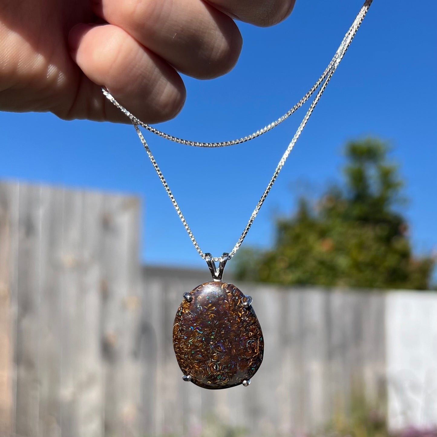 A Koroit boulder opal necklace dangling from a hand in natural sunlight, highlighting its earthy ironstone matrix and flashes of blue fire.