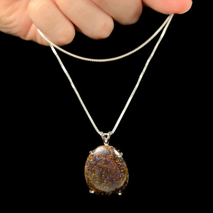 A Koroit boulder opal necklace dangling from a hand, displaying its earthy ironstone matrix and flashes of blue fire against a black background.