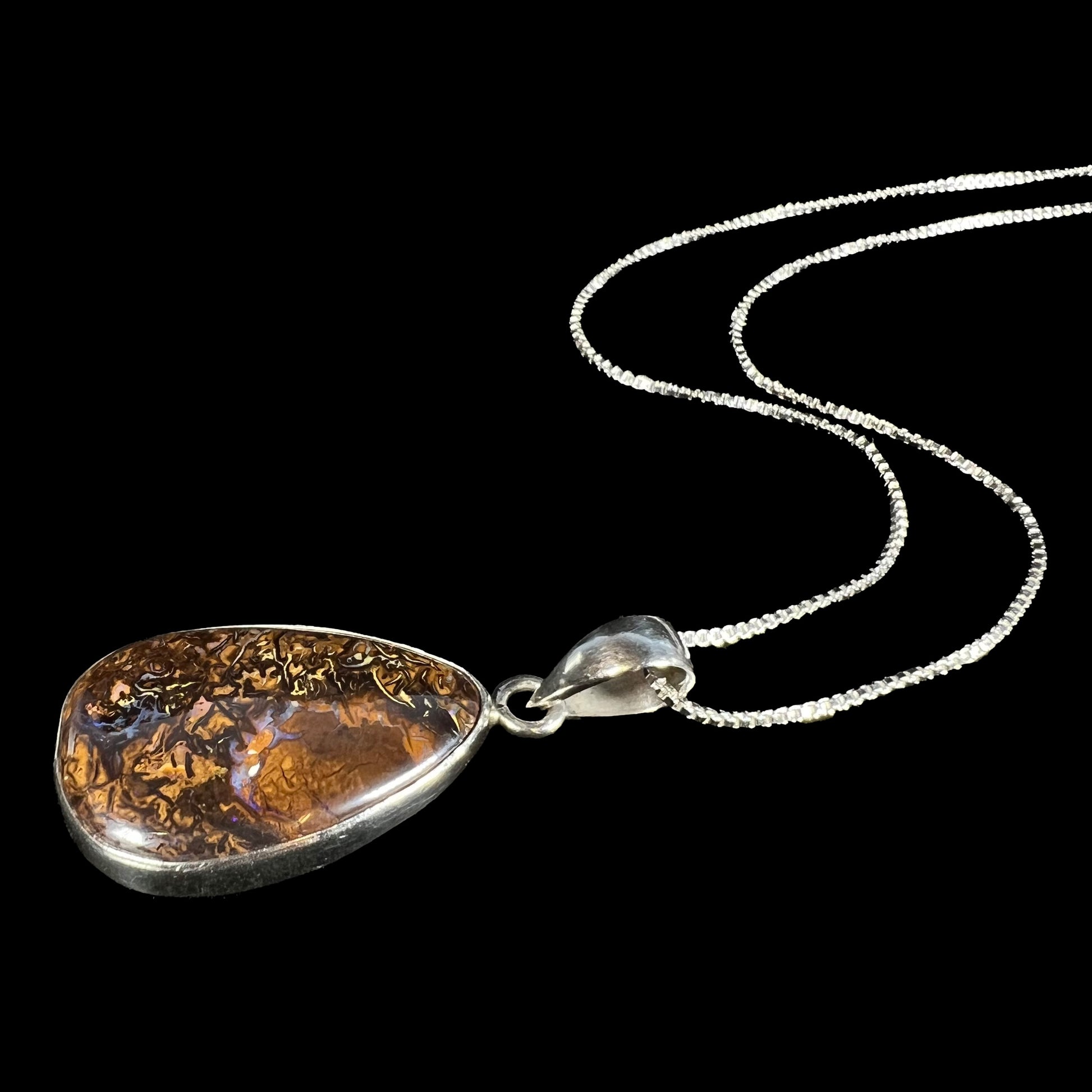 A pear shaped Koroit boulder opal necklace in sterling silver shown from an angle laying against a black backdrop.