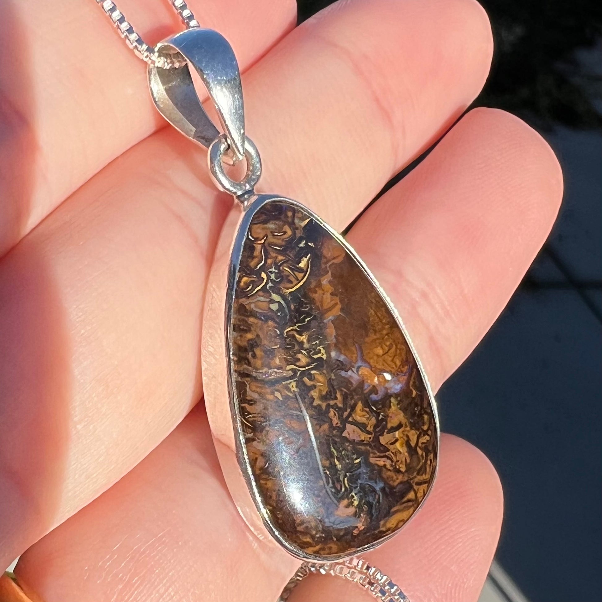 A pear shaped Koroit boulder opal pendant in sterling silver shown close-up in a hand in the sunlight from an angle.