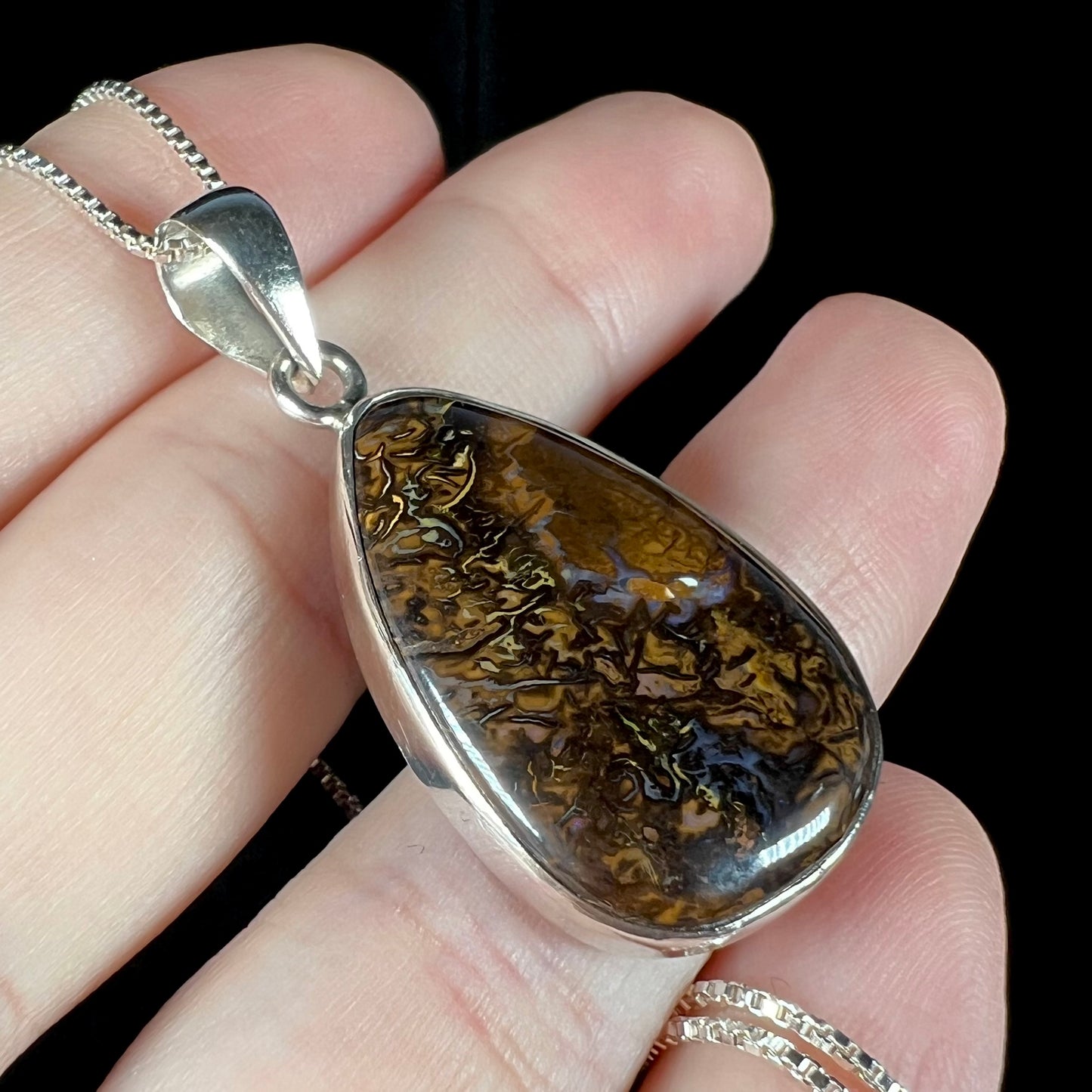 A pear shaped Koroit boulder opal pendant in sterling silver shown close-up in a hand from an angle.