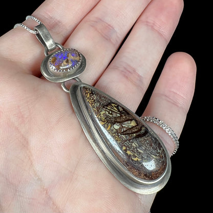 Angled close-up of a Southwest-style silver koroit boulder opal pendant held in a hand.  The pear shaped opal has tribal designs.