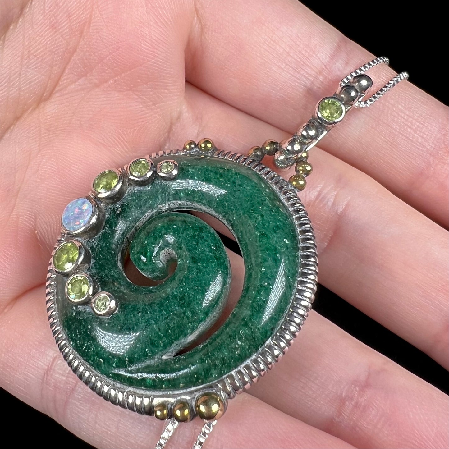 A men's aventurine koru necklace, accented with opal and peridot, shown from an angle in a hand.