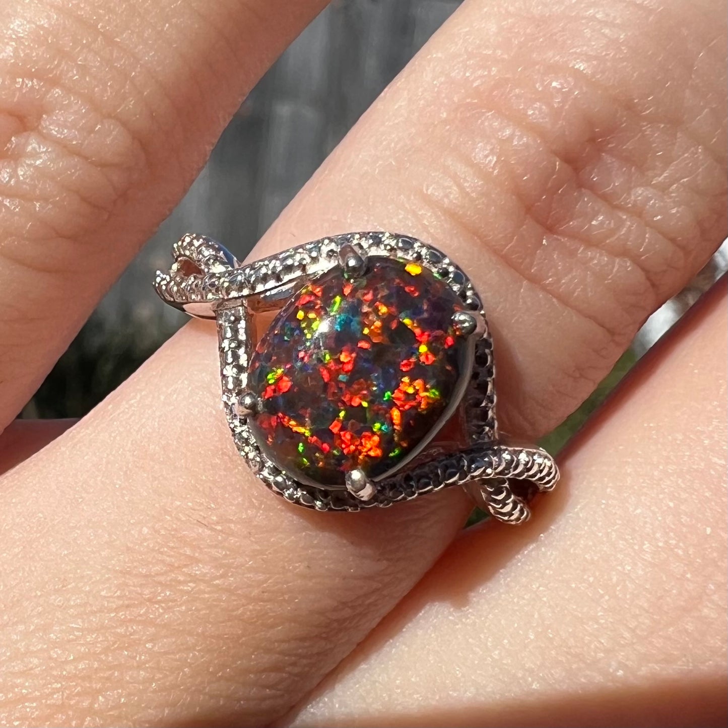 A lab-created black opal solitaire ring with red color play in sterling silver, shown close-up on a finger in sunlight.