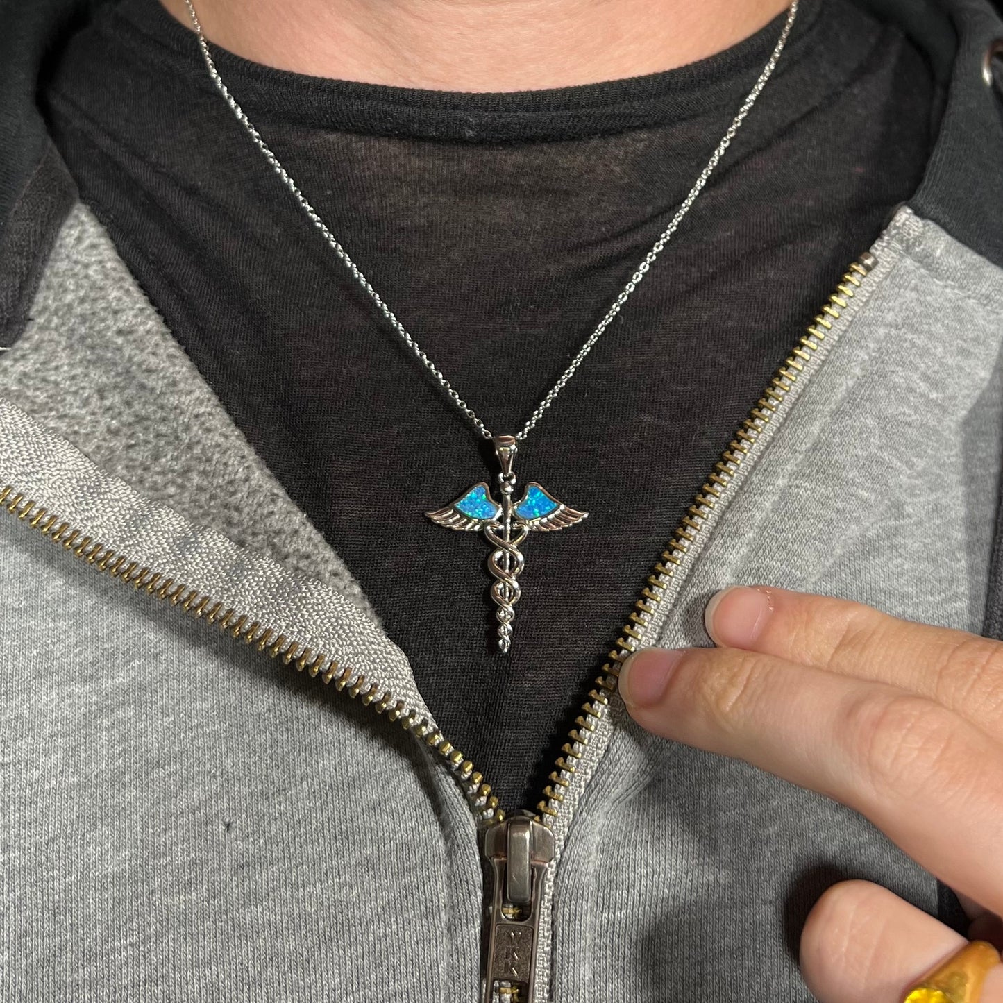 A silver plated medical caduceus necklace with blue lab-created opal inlay worn around a neck.