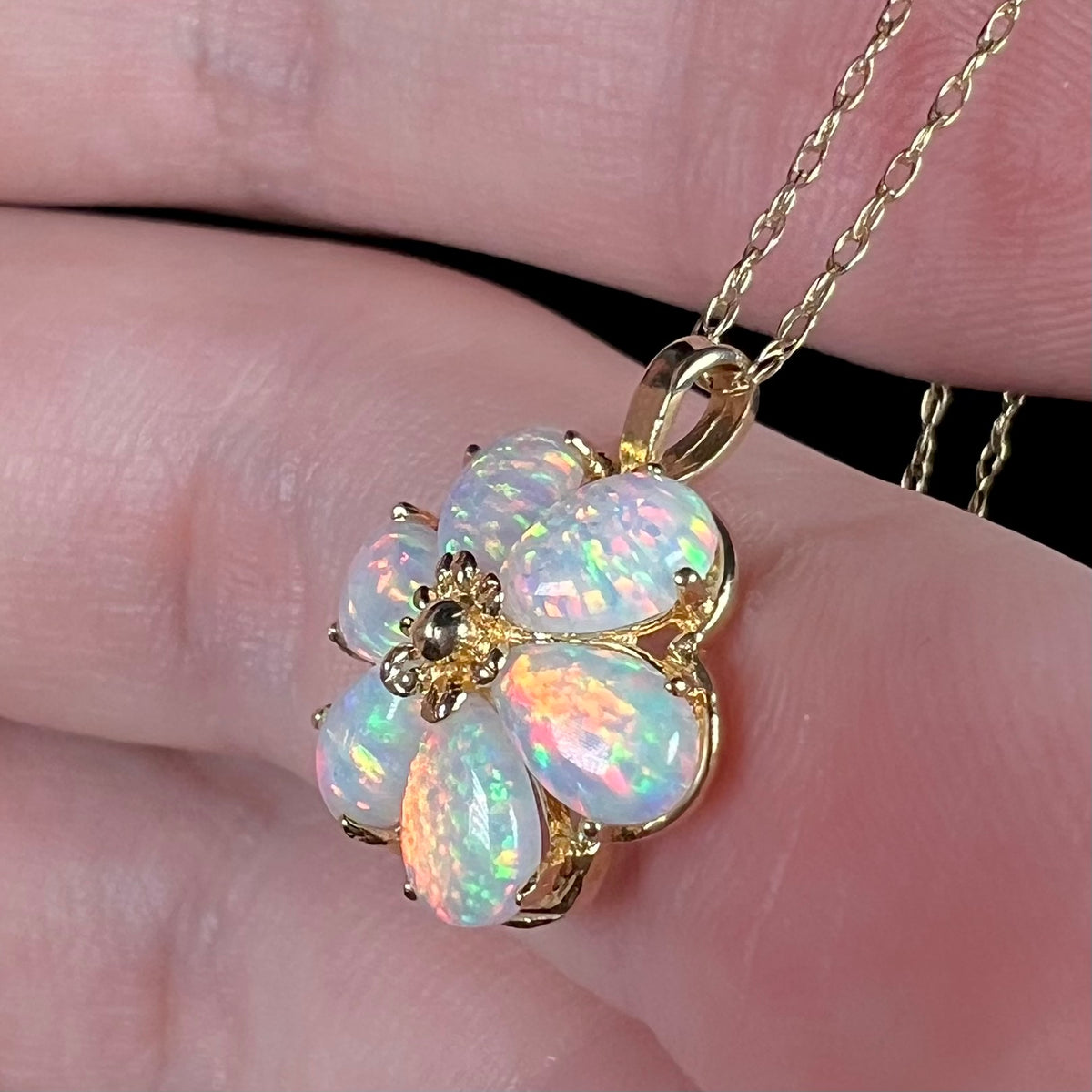 A lab-created opal flower pendant in 14k gold on a dainty gold chain, shown from the side view close-up in a hand