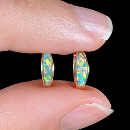 Close-up of a pair of yellow gold lab-created opal inlay huggie hoop earrings held between two fingers against a black background.