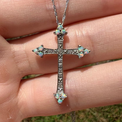 Close-up front view of a sterling silver lab-created opal cross necklace with CZ accents held in sunlight.