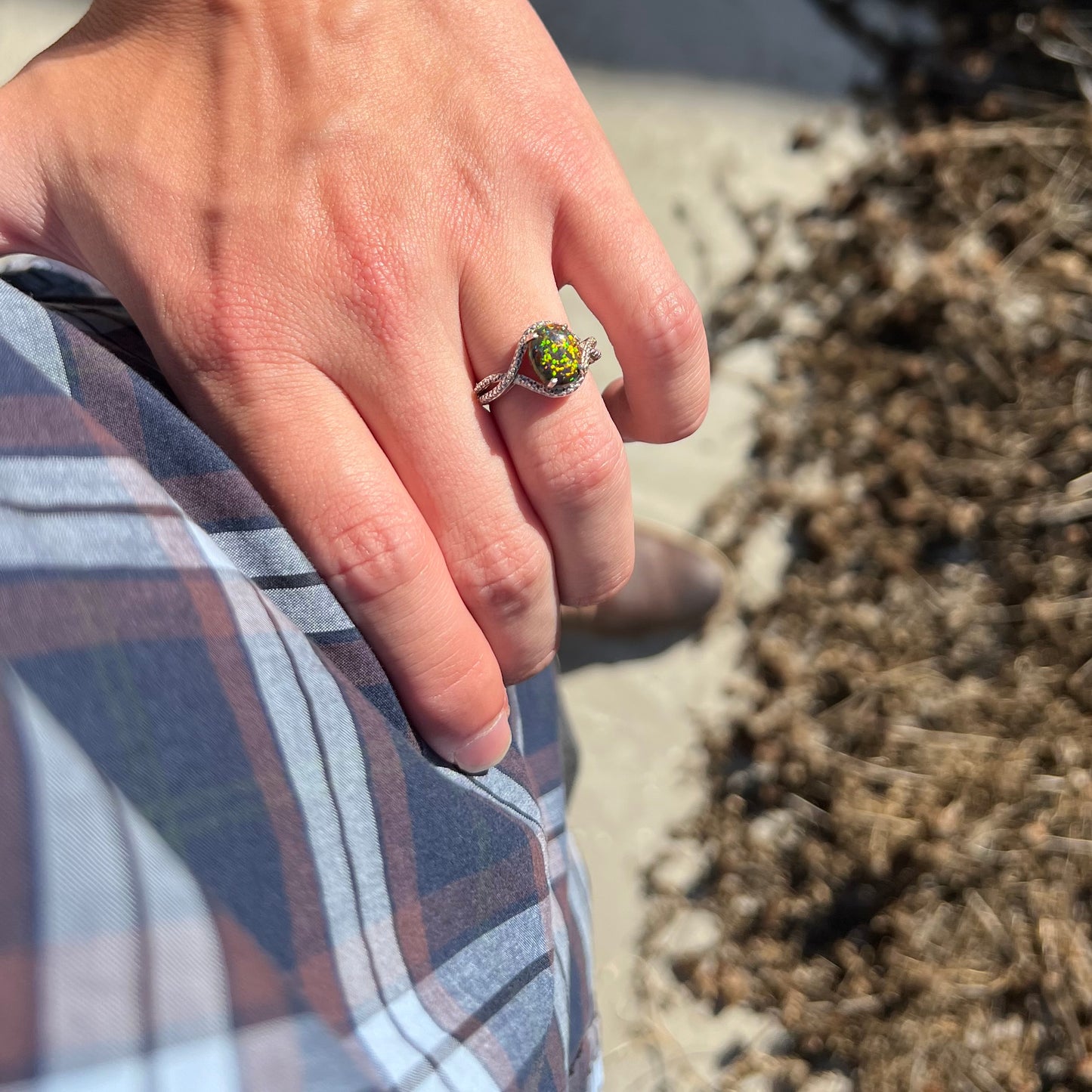 A lab-created black opal solitaire ring with green color play in sterling silver, worn on a hand resting on the model's hip.
