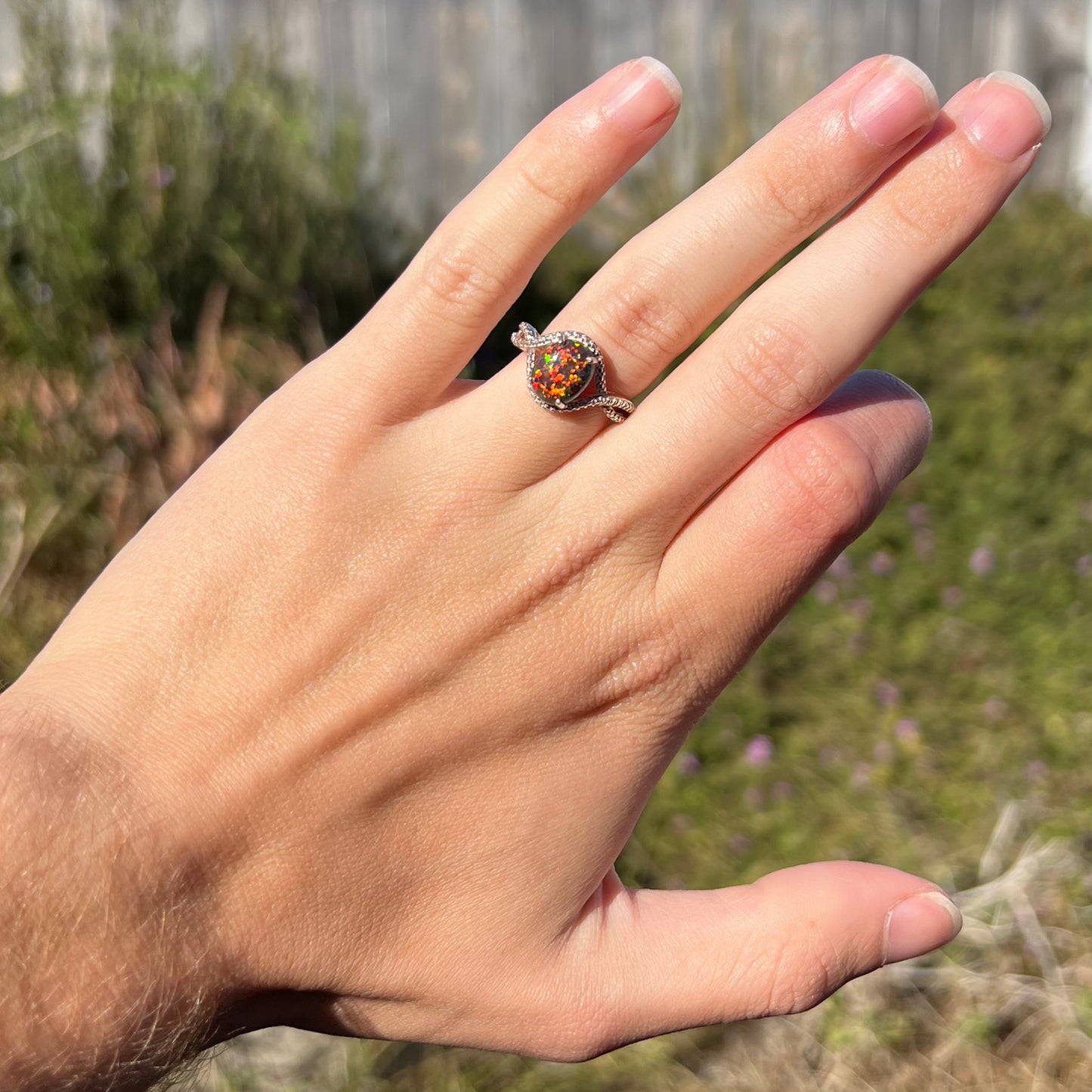 A lab-created black opal solitaire ring with red color play in sterling silver, shown on a hand in sunlight.