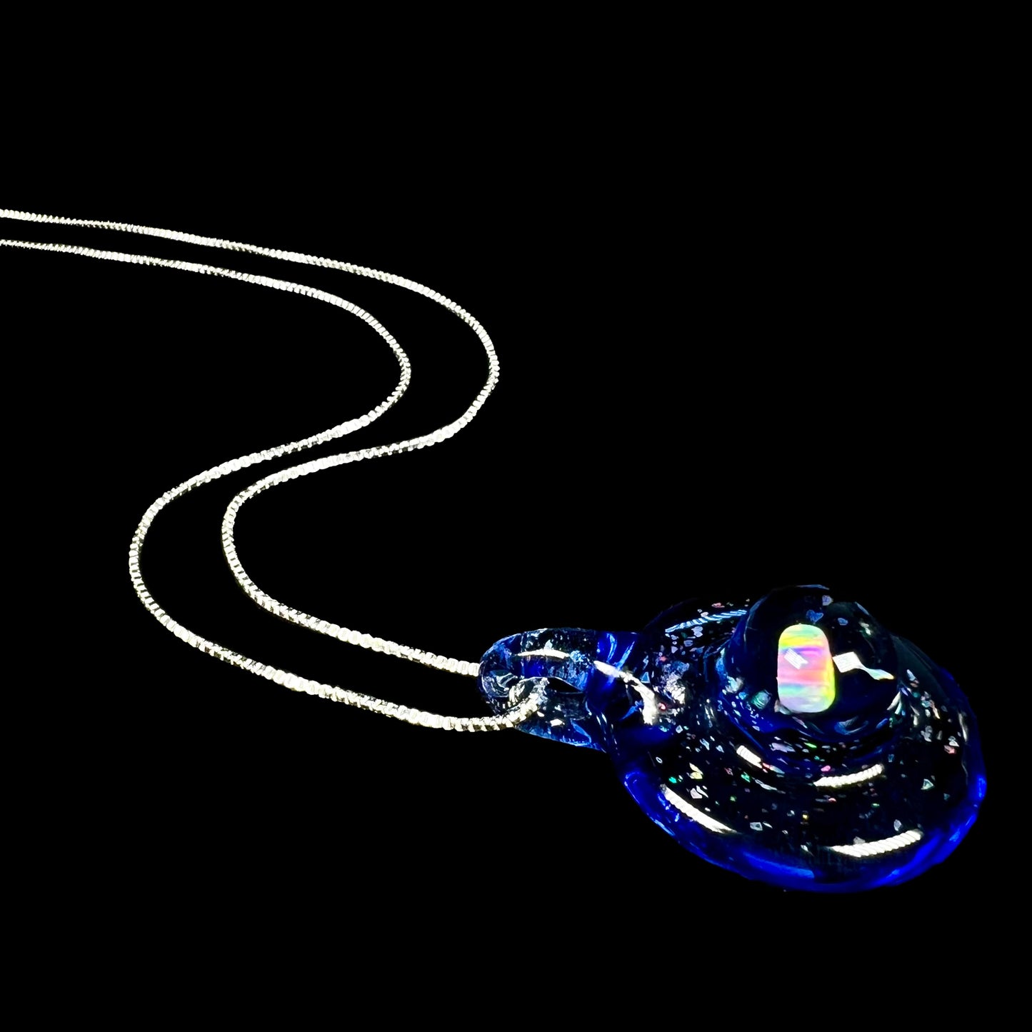 A hand-blown lab opal in blue glass necklace on a silver chain, shown from an angle laying against a black background.
