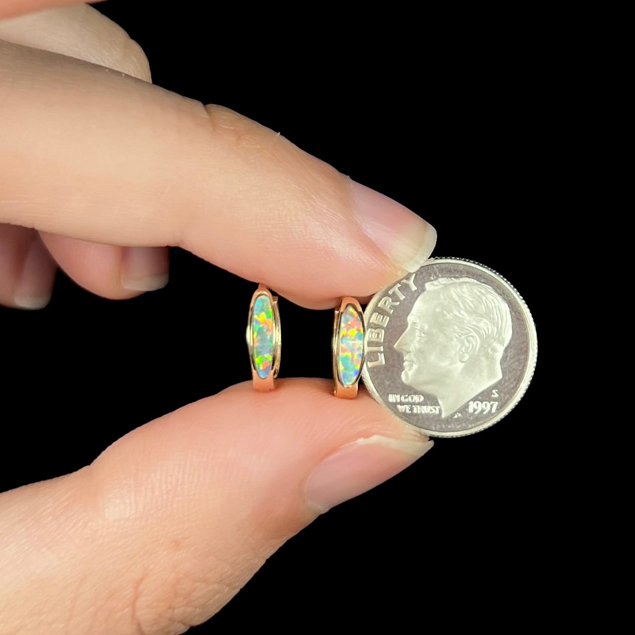 A pair of yellow gold lab-created opal inlay huggie hoop earrings held between two fingers with a dime coin.