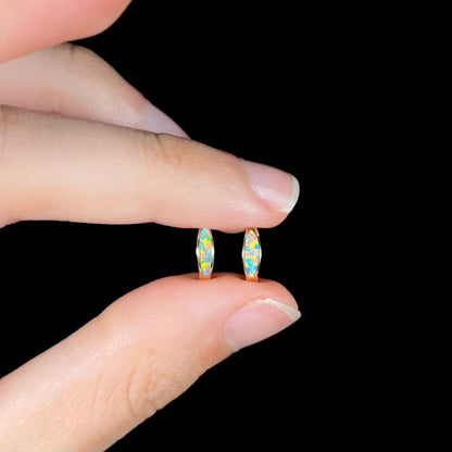 A pair of yellow gold lab-created opal inlay huggie hoop earrings held between two fingers against a black backdrop.